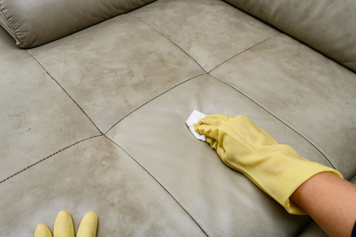 Nahaufnahme einer Hand mit gelbem Handschuh, die sorgfältig mit einem weißen Tuch über die Oberfläche eines braunen Ledersofas wischt, um es zu reinigen und zu pflegen